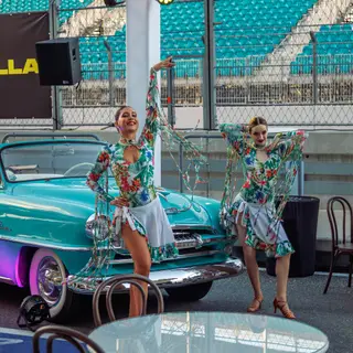 Performers in tropical costume beside a vintage car in front of the Autodrome grandstand