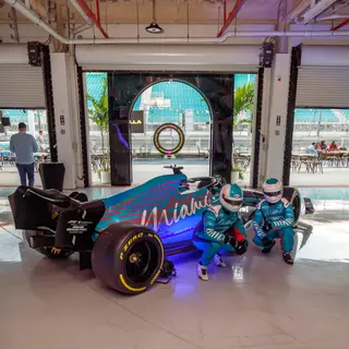 Race-suited crew beside an F1 show car in the pit garage at Miami International Autodrome