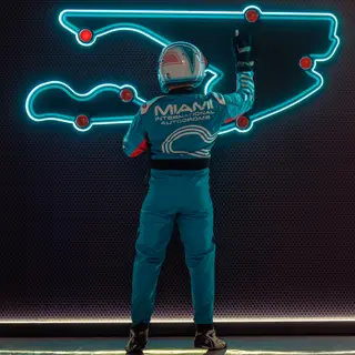 Race-suited crew member in front of a neon outline of the Miami International Autodrome circuit