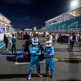 Two race-suited SimsForHire crew on the pit straight at night with guests and grandstands behind